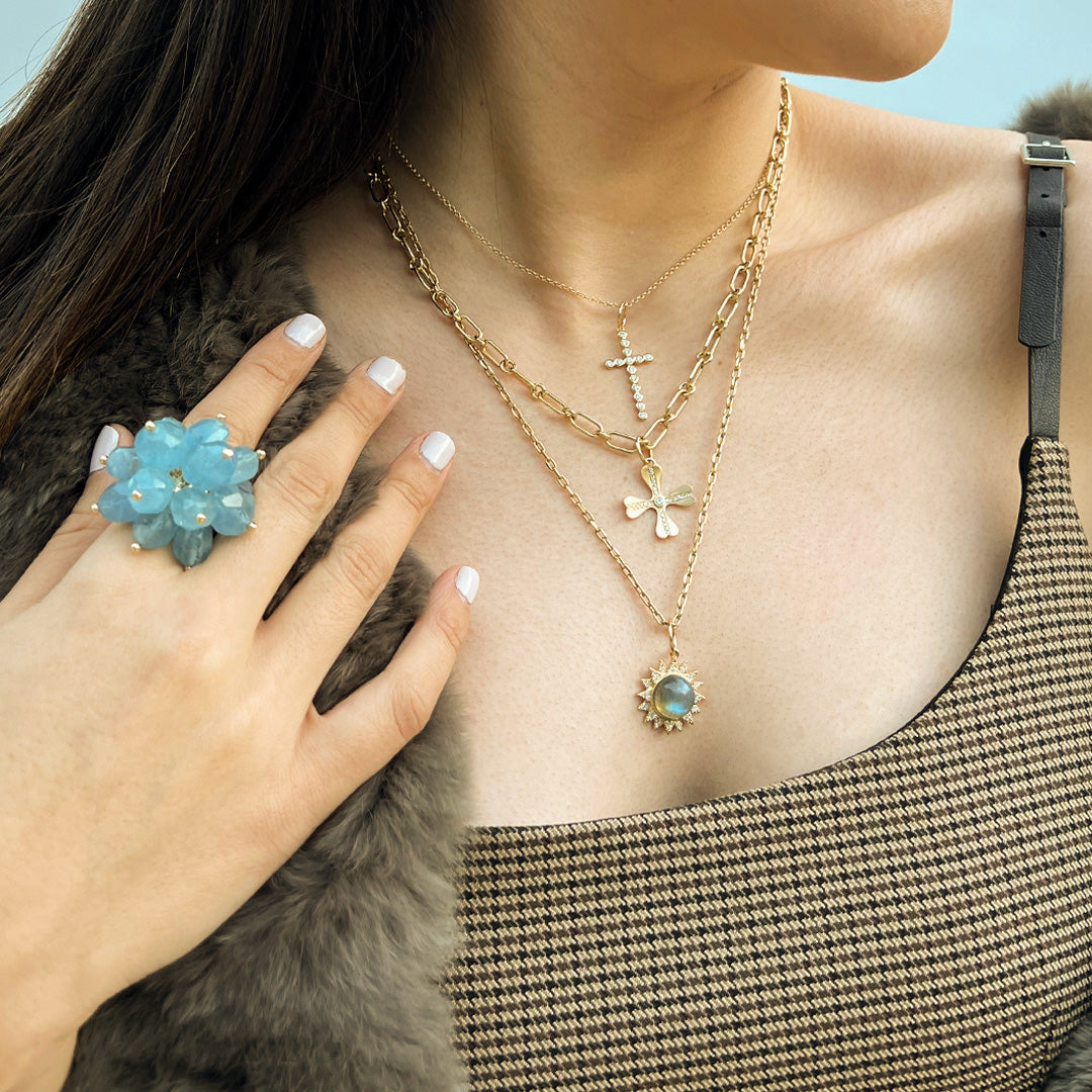 Woman is wearing the Aquamarine Pom Pom Ring, the Bezel-set Diamond Cross Necklace, the Floral Necklace, and the Labradorite Sunburst Necklace
