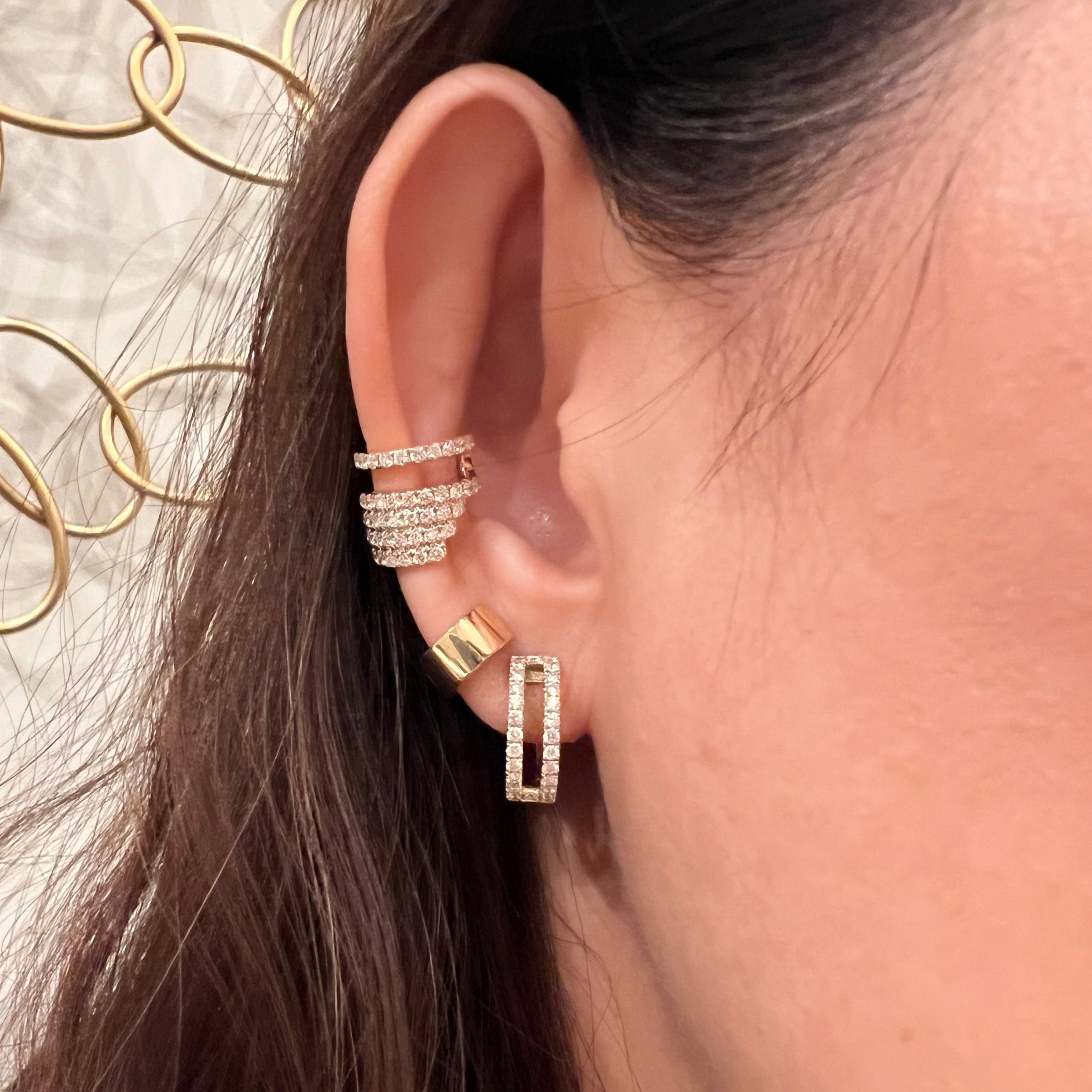Close-up of an ear wearing multiple 14K gold and diamond earrings (Chandelier Ear Cuff, Chubby Huggies, and Luxe Open Bar Diamond Huggies) with a blurred background.