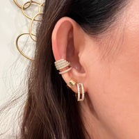 Close-up of an ear wearing multiple 14K gold and diamond earrings (Chandelier Ear Cuff, Chubby Huggies, and Luxe Open Bar Diamond Huggies) with a blurred background.