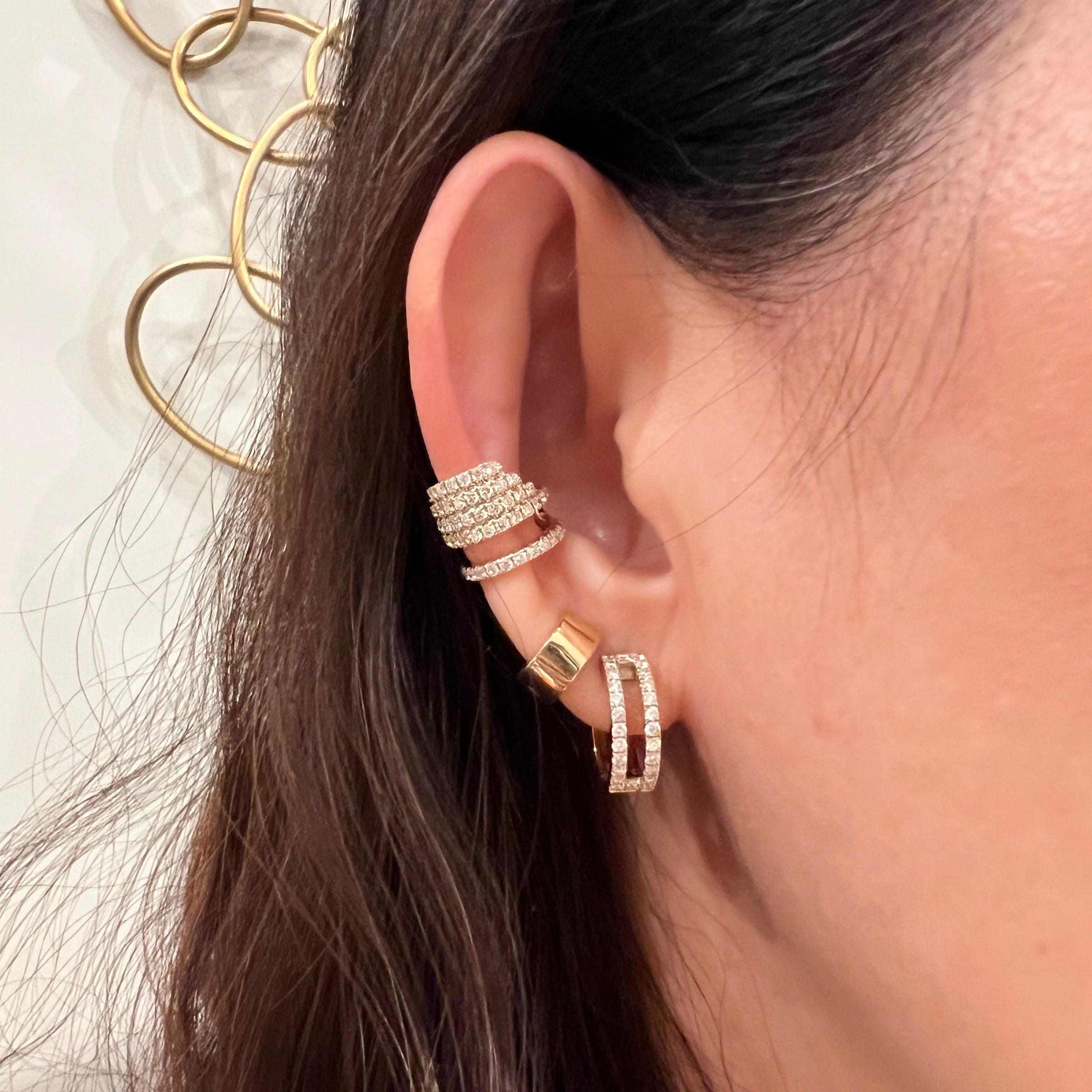 Close-up of an ear wearing multiple 14K gold and diamond earrings (Chandelier Ear Cuff, Chubby Huggies, and Luxe Open Bar Diamond Huggies) with a blurred background.