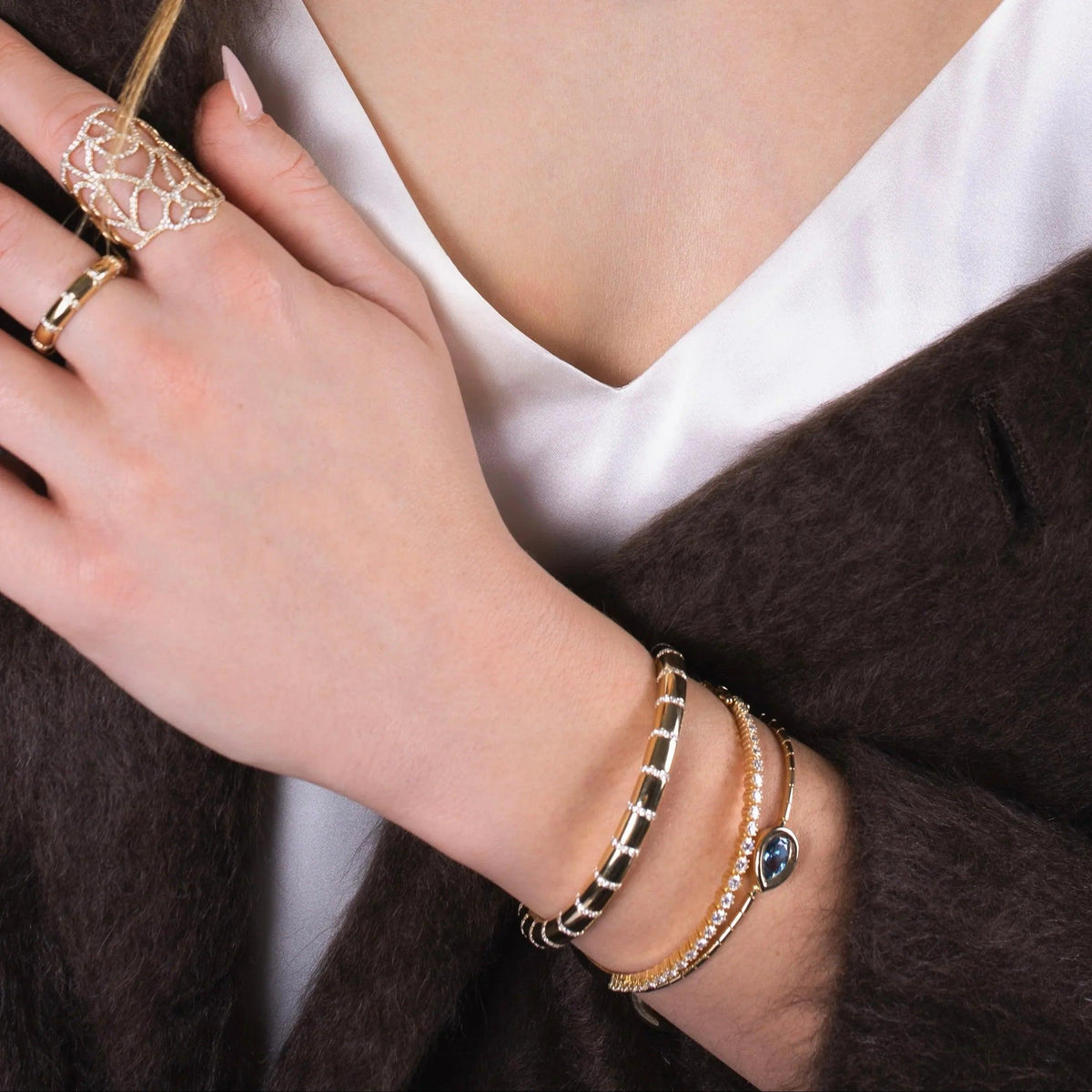 Close-up of a person wearing London Blue Topaz Liquid Teardrop Bracelet, Diamond Section Stretch Bracelet, 3mm Diamond Stretch Tennis Bracelet with a white shirt and brown coat.