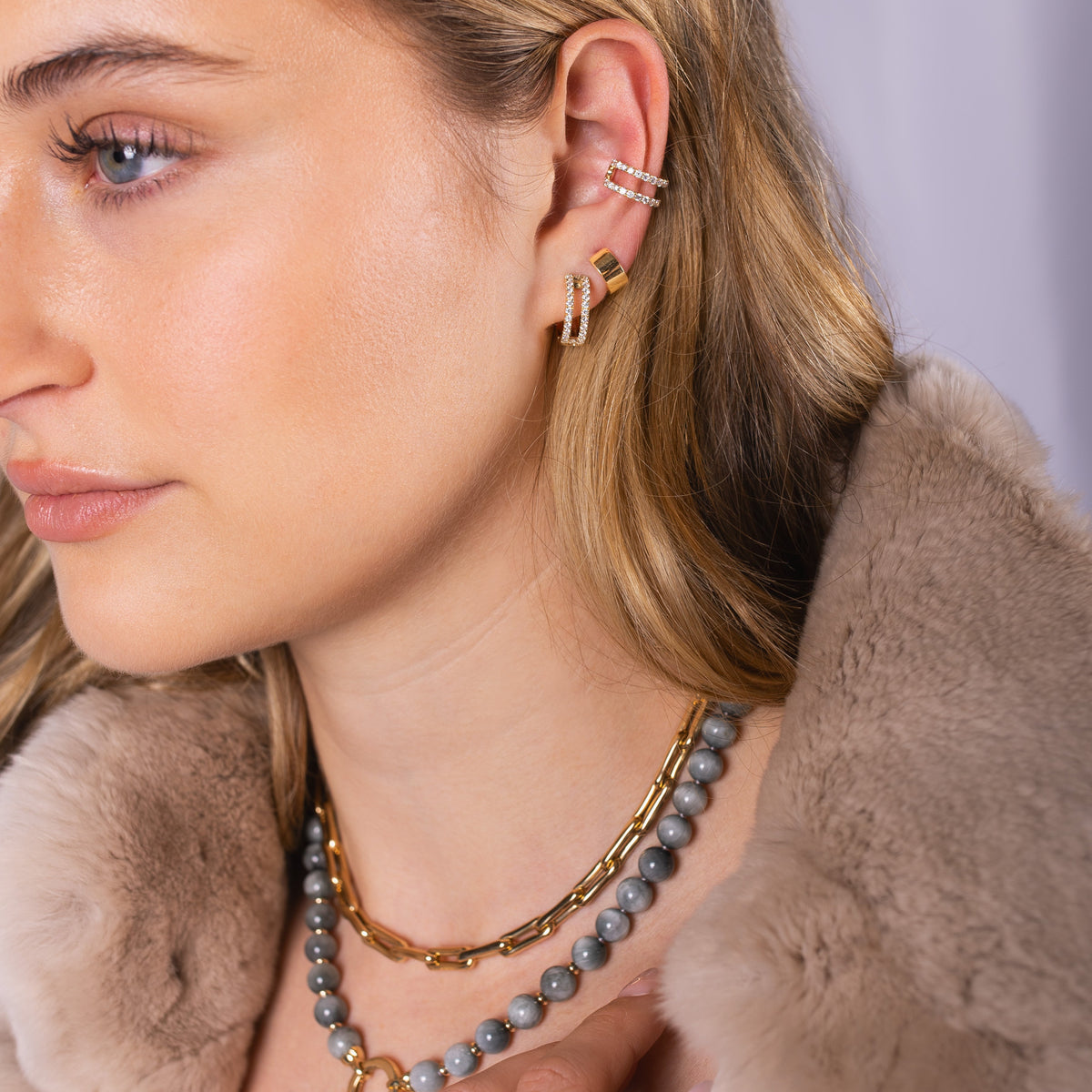 Close-up of a woman wearing Luxe Open Bar Diamond Huggies, the Chubby Huggies, the Open Bar Diamond Ear Cuff and the Bone Link Chain with a fur coat.