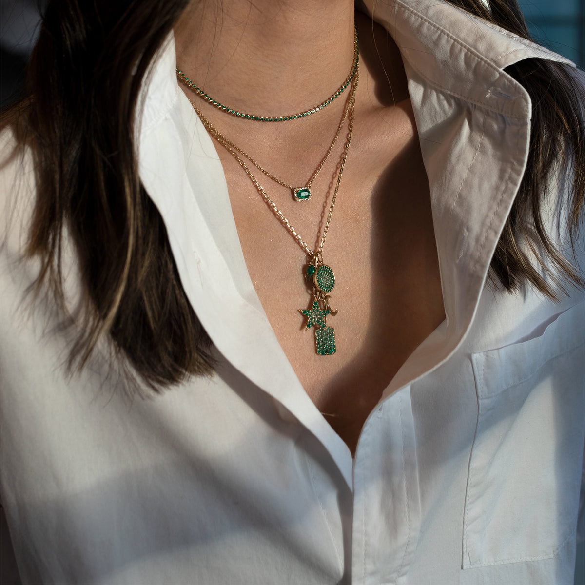 Close up of a person wearing the Emerald Necklace, the Emerald Tennis Necklace, and a 5 emerald charm necklace